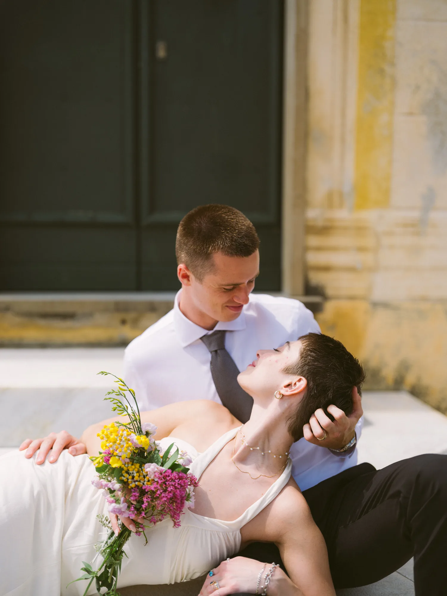 A groom in a white shirt and black tie is holding a bride's face, both lying on a stone surface.