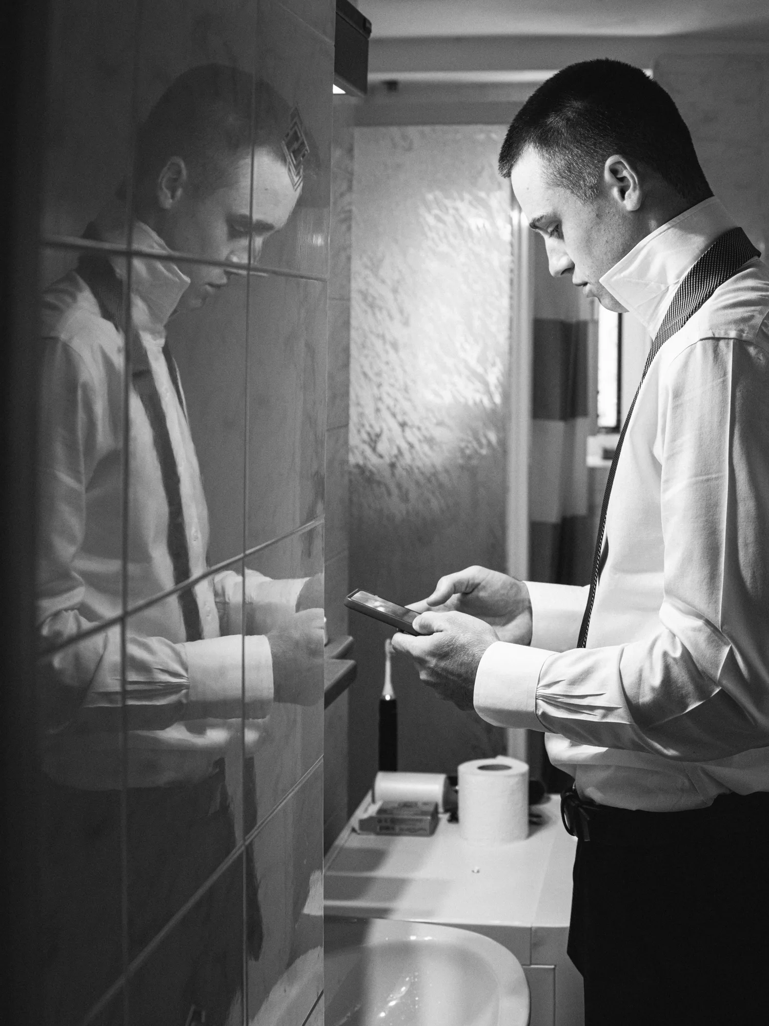 A groom in a white shirt and black tie is standing in a bathroom, looking at his phone.