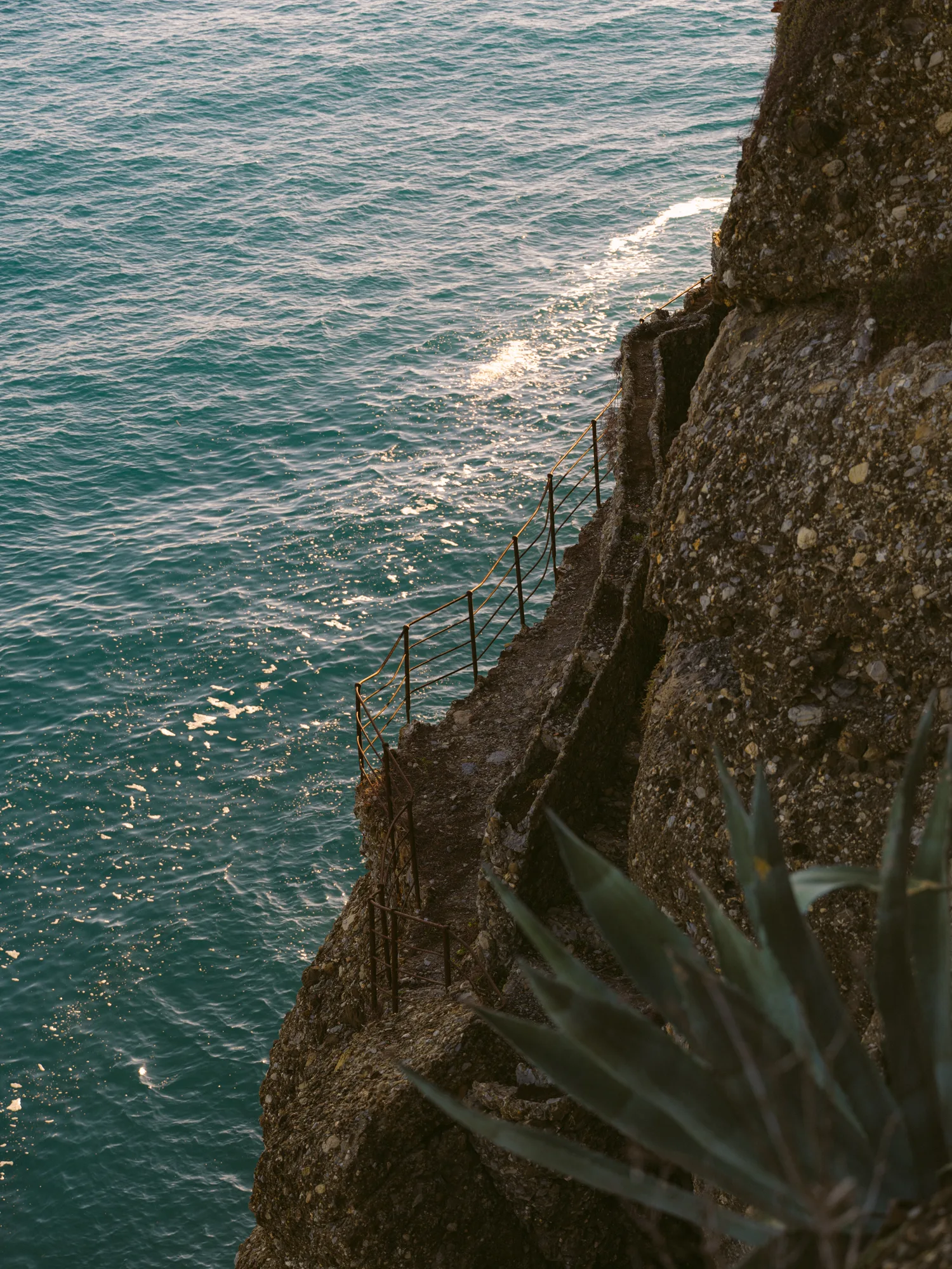 The photograph shows a rocky cliffside with a metal railing along the edge, overlooking a body of turquoise blue water.