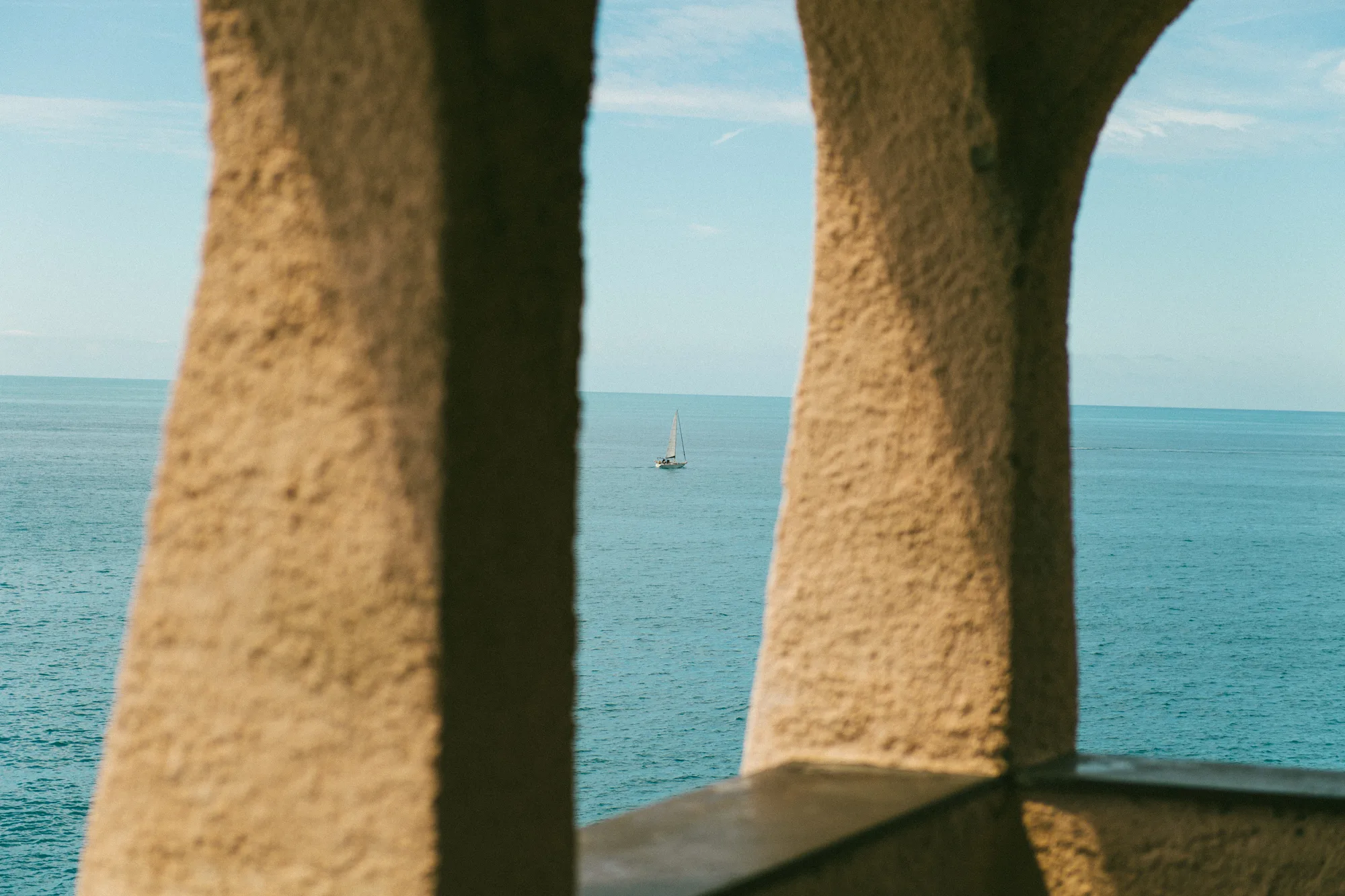 A sailboat is sailing on the calm blue sea, framed by a stone archway.