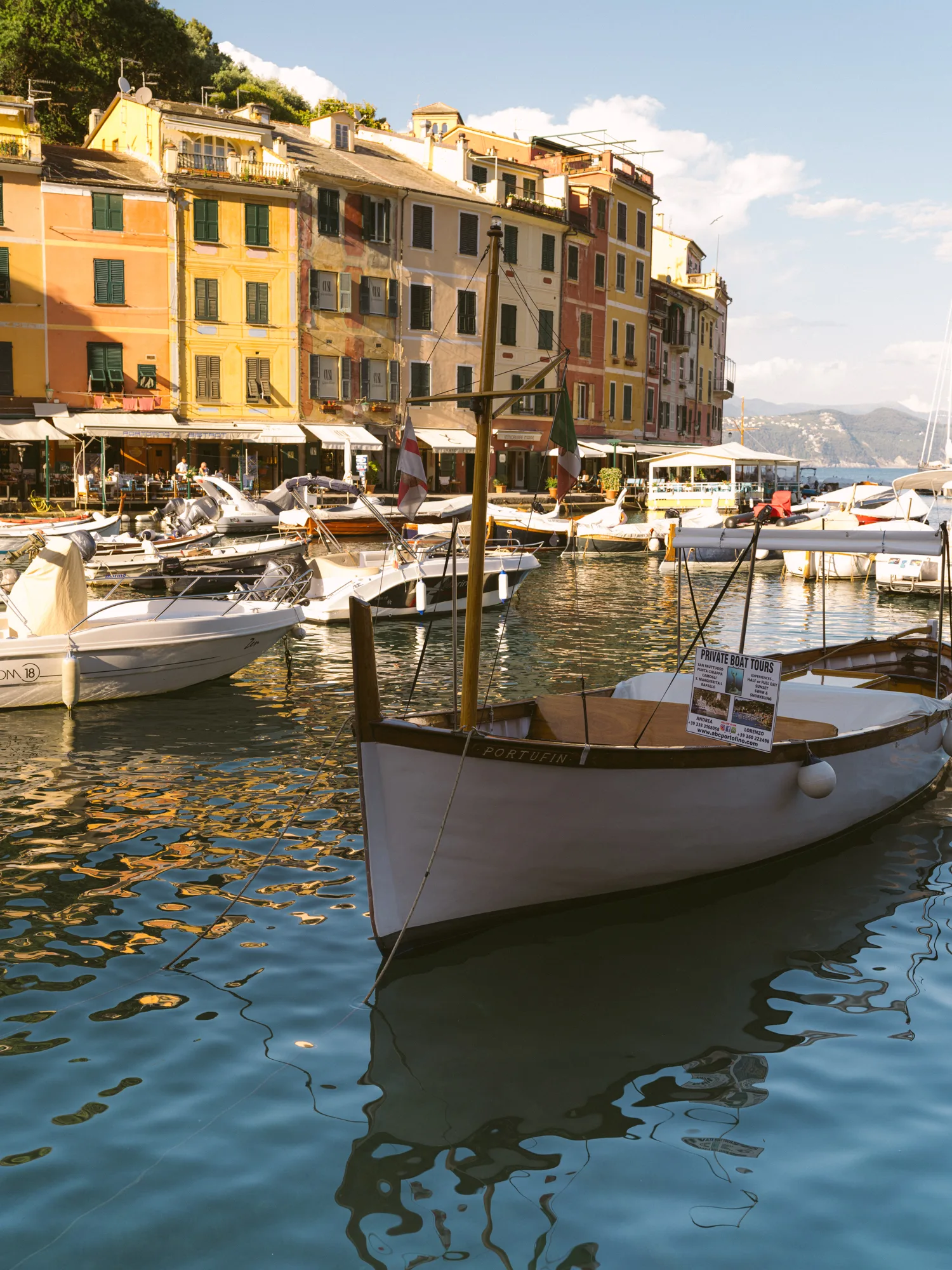 The photograph shows a serene harbor with several boats docked, including a white boat with a sign reading "PRIVATE BOAT TOURS." The background features colorful buildings with balconies and shutters, and a clear blue sky with a few clouds in Portofino