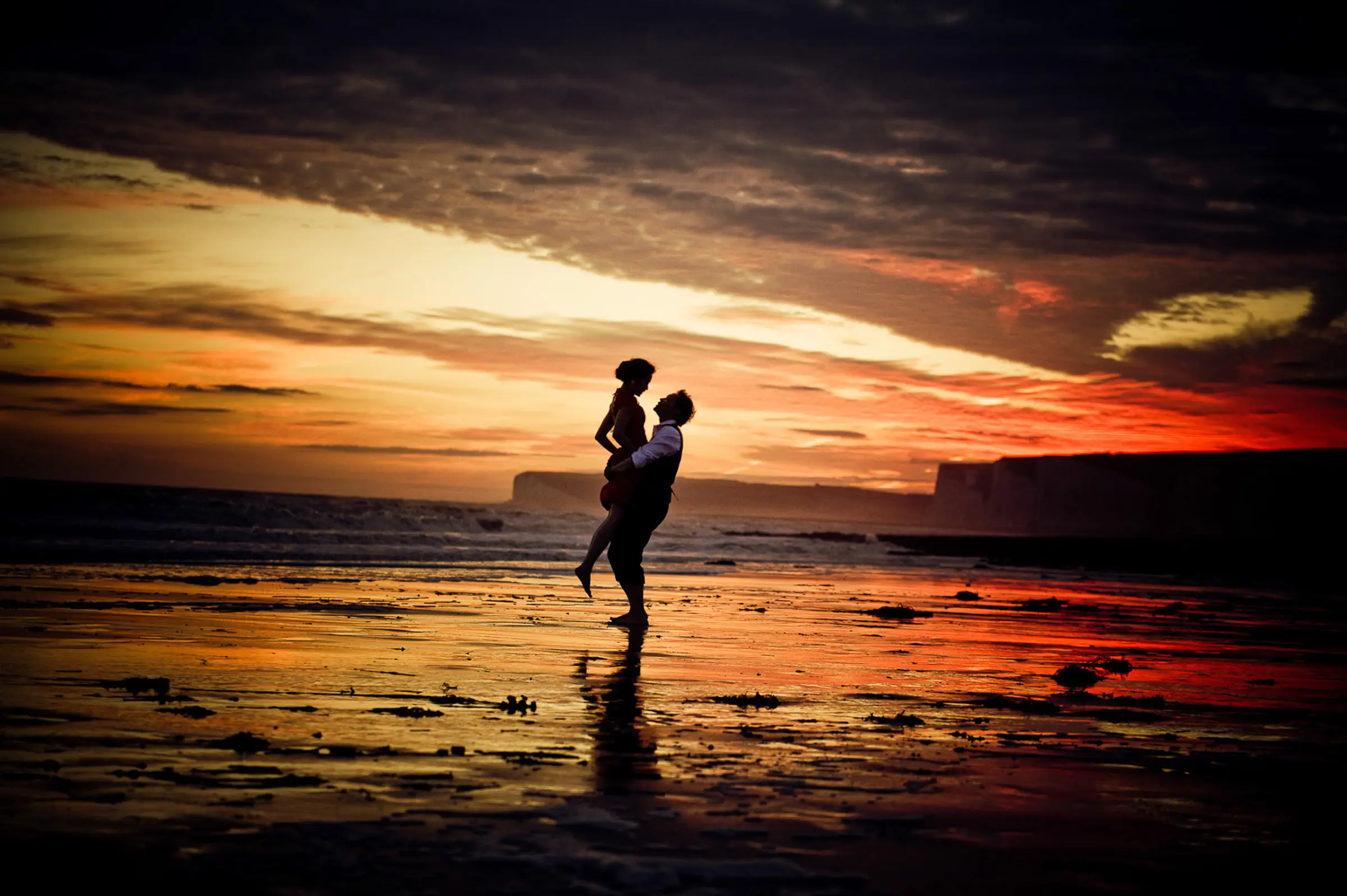 A couple embrace on a beach, silhouetted against an orange and gold sunset