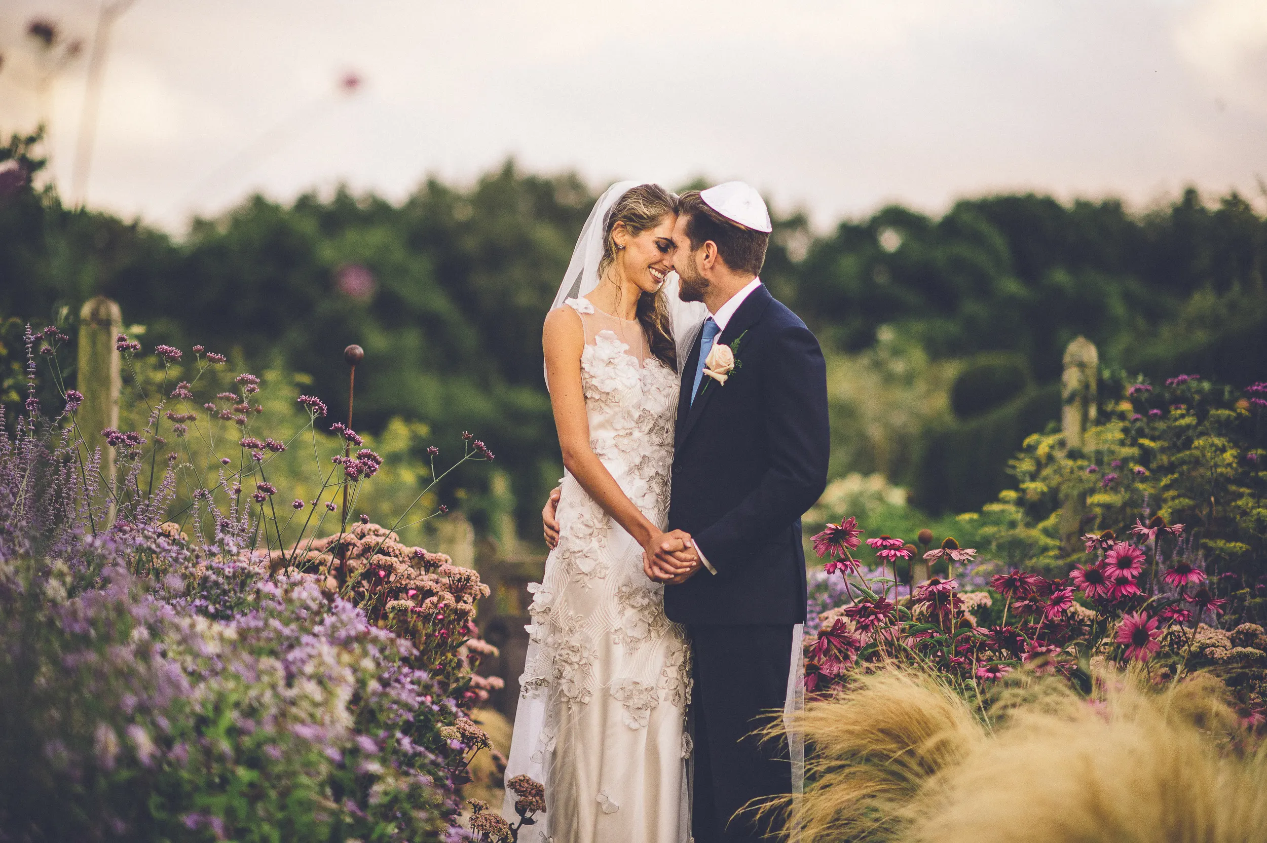 Bride and groom standing in a garden, holding hands and smiling, surrounded by colorful flowers and greenery by Hampshire wedding photographer