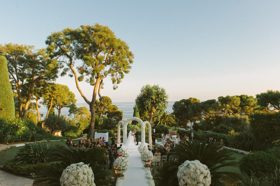 Bride and groom during a luxury outdoor ceremony at Grand-Hôtel du Cap-Ferrat, surrounded by floral arches, guests, and Mediterranean views.
